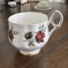 A white porcelain teacup with gold trim and red rose designs sits beside a Woulind Chile Serving Dish on a wooden table, with decorative photo frames and a candle softly blurred in the background.