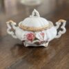 The Woulind Chile Serving Dish is a white porcelain sugar bowl with ornate gold-trimmed handles, a floral lid, elegant curved feet, and a gently worn gold rim, adding charm on a wooden table with a softly blurred background.