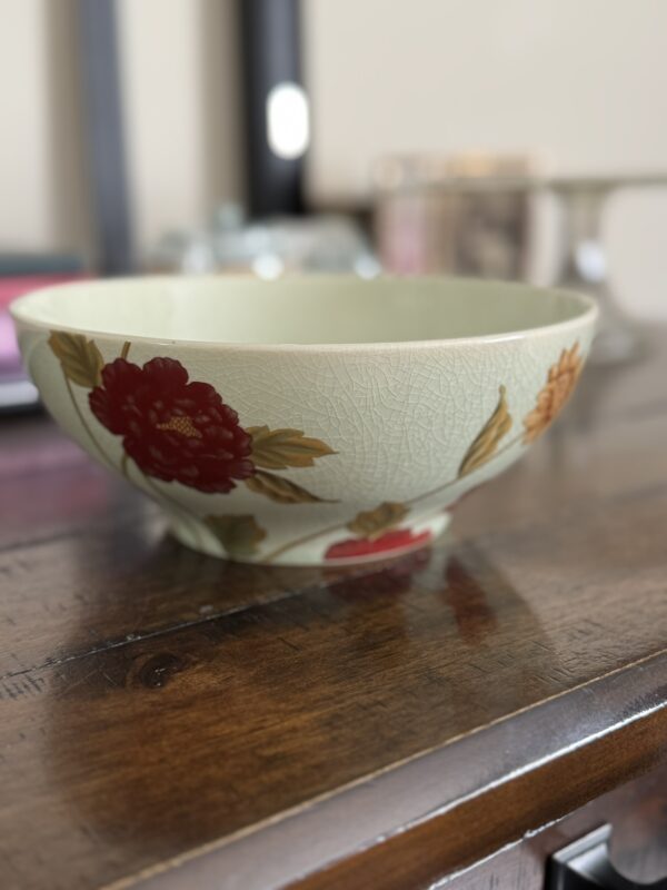 The Woulind Chile Serving Dish, featuring a crackled light green ceramic surface with painted red and yellow flowers, rests on a dark wooden table. A softly blurred background reveals household items bathed in gentle natural light.