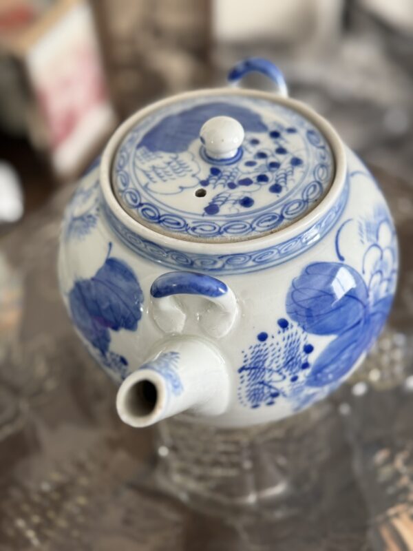 A white porcelain teapot with blue floral and bird patterns sits on a glass surface beside the Woulind Chile Serving Dish. The teapot features a round body, curved spout, handle, and lid with knob against a softly blurred background.