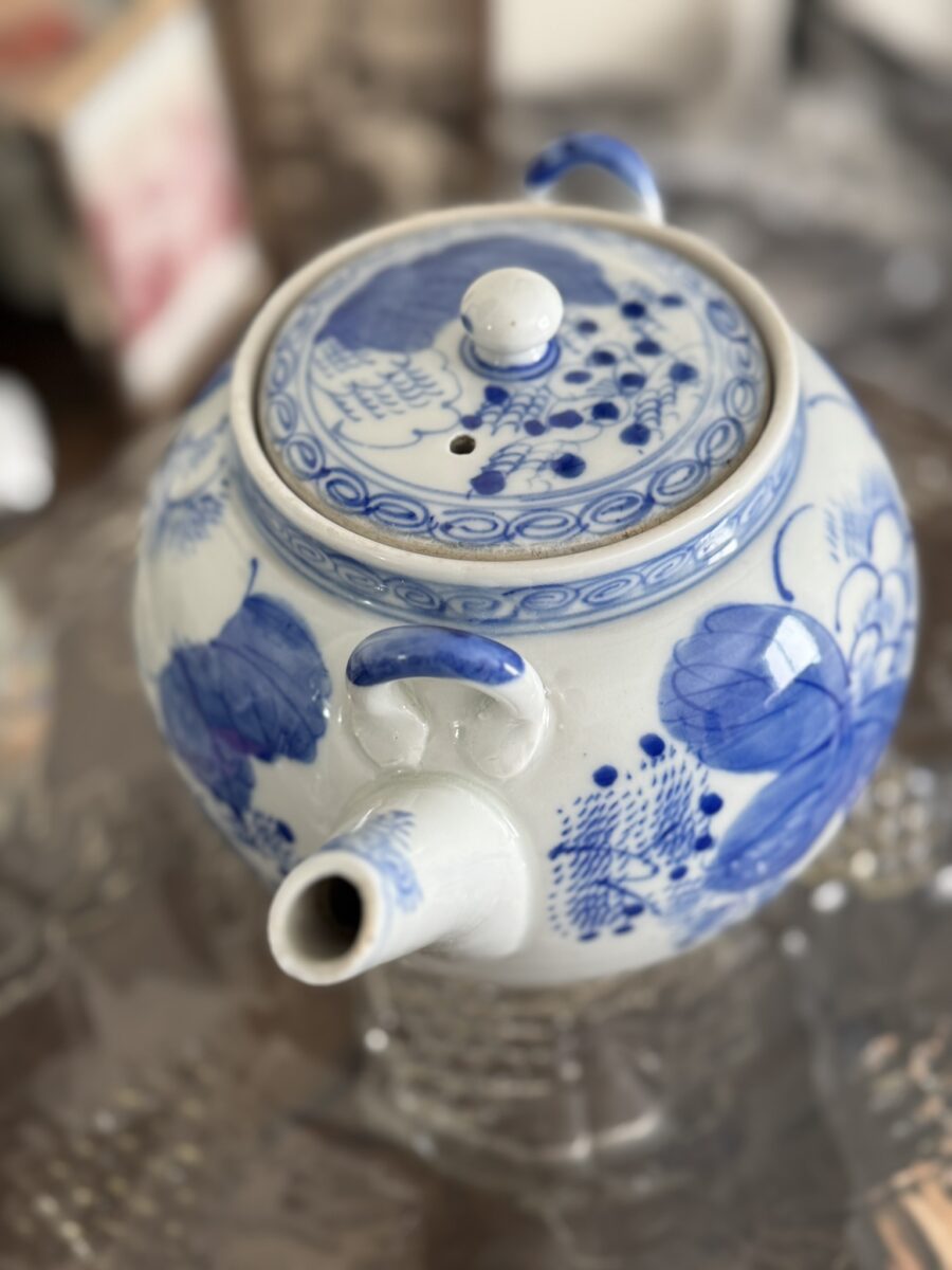 A white porcelain teapot with blue floral and bird patterns sits on a glass surface beside the Woulind Chile Serving Dish. The teapot features a round body, curved spout, handle, and lid with knob against a softly blurred background.