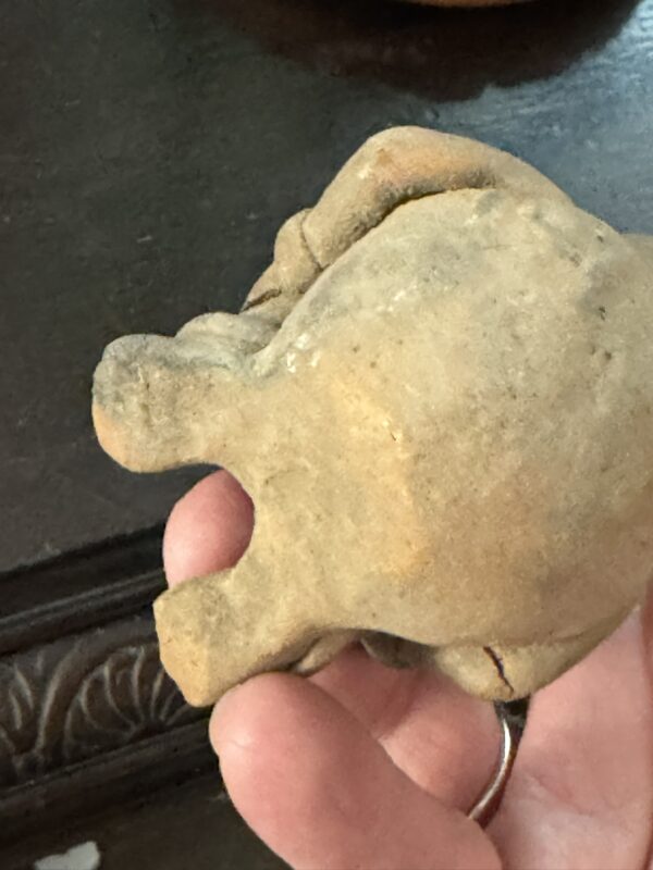 A hand holds the Mayan Figurine, a small, roughly textured tan clay sculpture resembling a crouched animal, possibly a frog or toad, displayed against a dark wooden surface with carved decorative patterns.