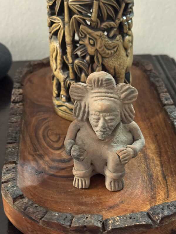 A Mayan Figurine with detailed facial features and pronounced hair stands on a carved wooden tray. Behind it is a cylindrical bamboo container with animal and foliage designs, set against a dark object and light wall.
