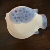 The Woulind Chile Serving Dish, shaped like a fish with blue painted details—eye, gills, fins, and dotted scales—sits on a dark wooden surface. Mostly white with blue accents, the ceramic dish has a slightly glossy finish.