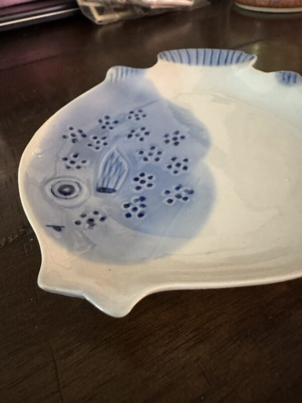The Woulind Chile Serving Dish, a ceramic dish shaped and painted like a fish in white with blue patterns and stylized details, rests on a dark wooden surface.