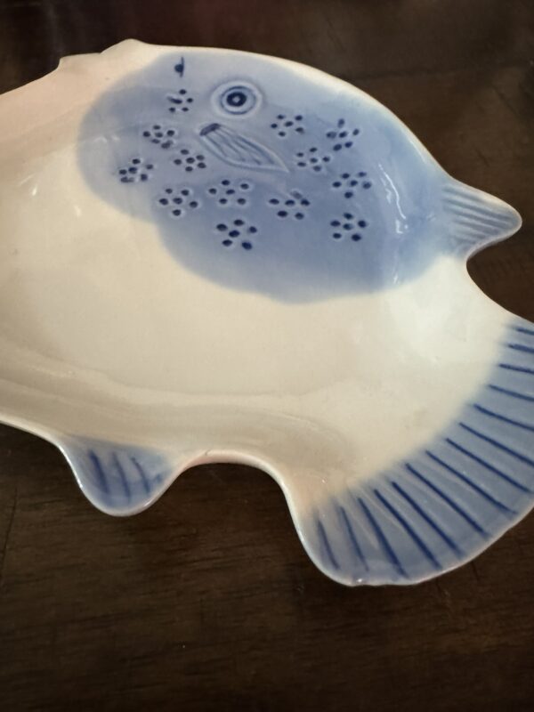 The Woulind Chile Serving Dish is a fish-shaped ceramic plate with blue detailing, featuring a stylized fish face, spots on the upper part, and blue lines on the tail and fins. It sits on a dark wooden surface.