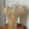 A translucent, beige glass vase with wavy, ruffled edges recalls the Woulind Chile Serving Dish. Its organic shape and glossy finish showcase hand-blown craftsmanship as it catches the light on a wooden surface.