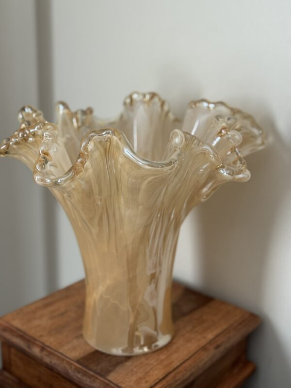 A translucent, beige glass vase with wavy, ruffled edges recalls the Woulind Chile Serving Dish. Its organic shape and glossy finish showcase hand-blown craftsmanship as it catches the light on a wooden surface.