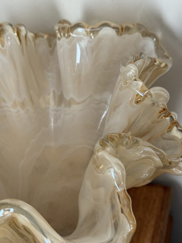 A close-up of the Woulind Chile Serving Dish, a cream-colored glass piece with ruffled, wavy edges and subtle gold accents. Its marbled patterns and flowing rim evoke organic shapes and shadows on a wooden surface.
