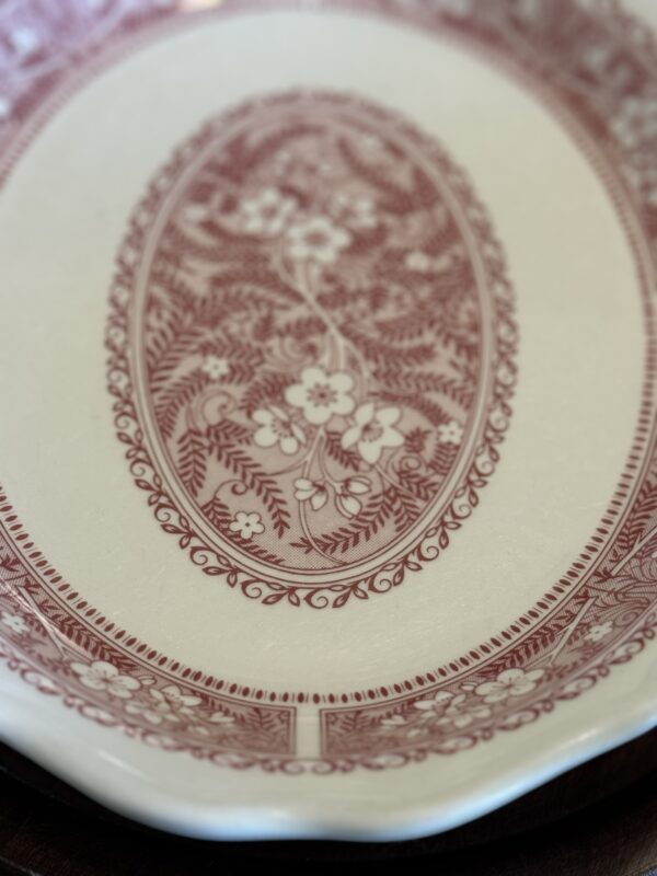 A close-up of the Woulind Chile Serving Dish, a white ceramic piece with a detailed maroon floral and foliage oval design at the center, ornate geometric and vine patterns on the rim, and subtle surface crazing.