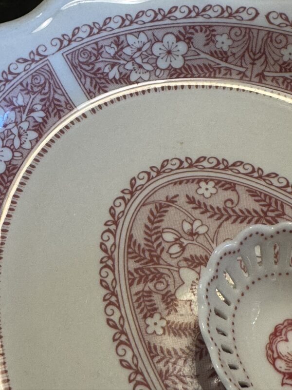 Close-up of two vintage ceramic plates with ornate red floral patterns on cream. The smaller plate is the Woulind Chile Serving Dish, featuring a scalloped, perforated edge and intricate overlapping details.