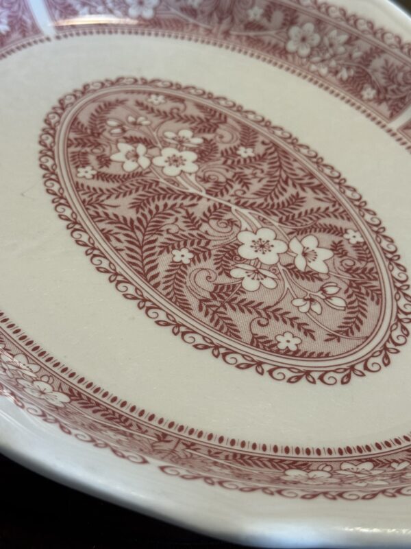 A close-up of the Woulind Chile Serving Dish, a white ceramic piece with a detailed red floral and leaf pattern in the center and rim, highlighted by swirling lines enclosed inside an oval with a bordered edge.