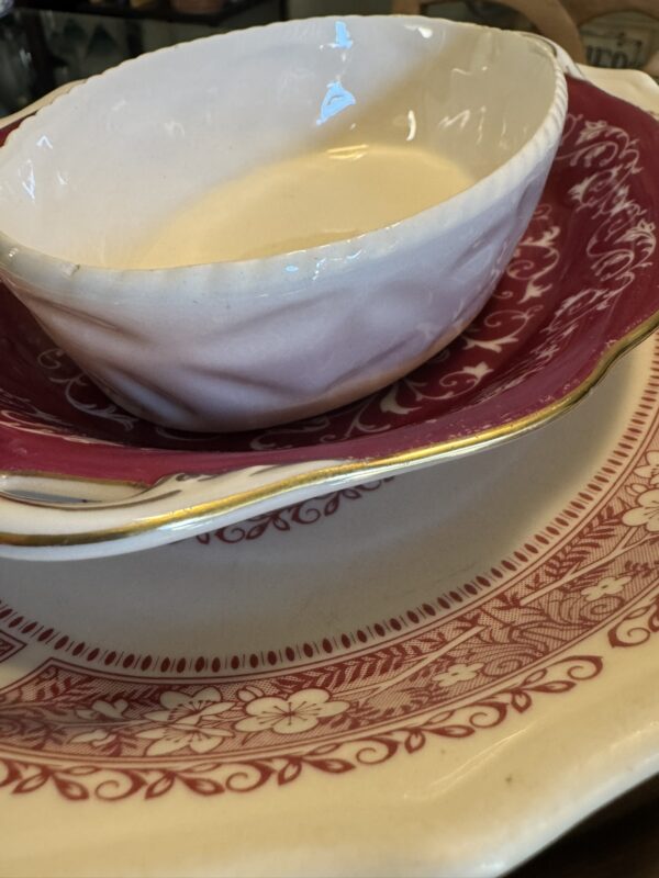 Close-up of three stacked dishes: a cream-colored bowl with a wavy texture sits atop the Woulind Chile Serving Dish, which features deep pink color, white floral patterns, and gold trim, all resting on a large cream plate with pink designs.