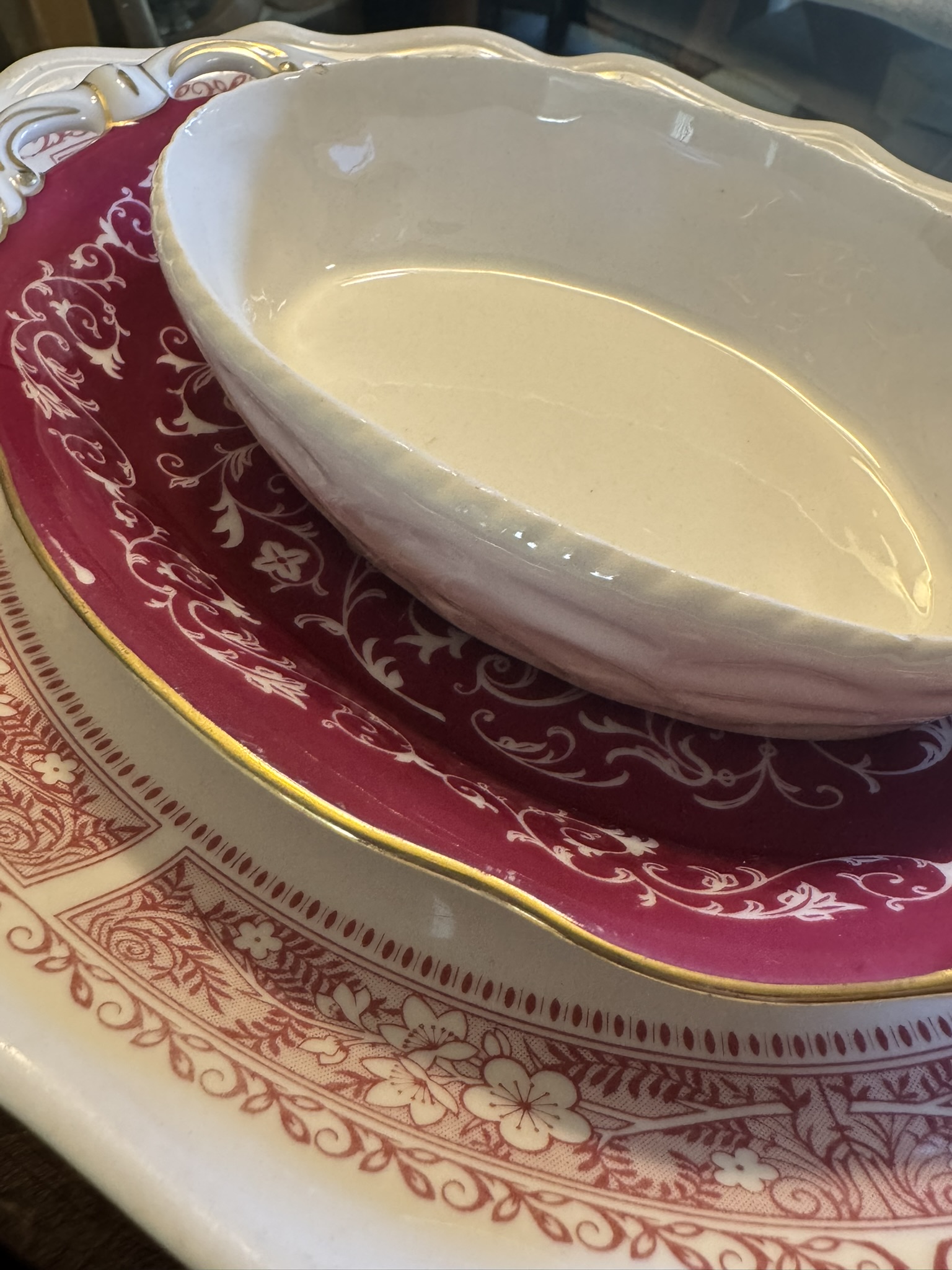 Three nested dishes: a Woulind Chile Serving Dish—white and oval—on top, a red porcelain plate with gold trim and white floral scrollwork in the middle, and a larger cream-colored plate with intricate red border patterns on the bottom.