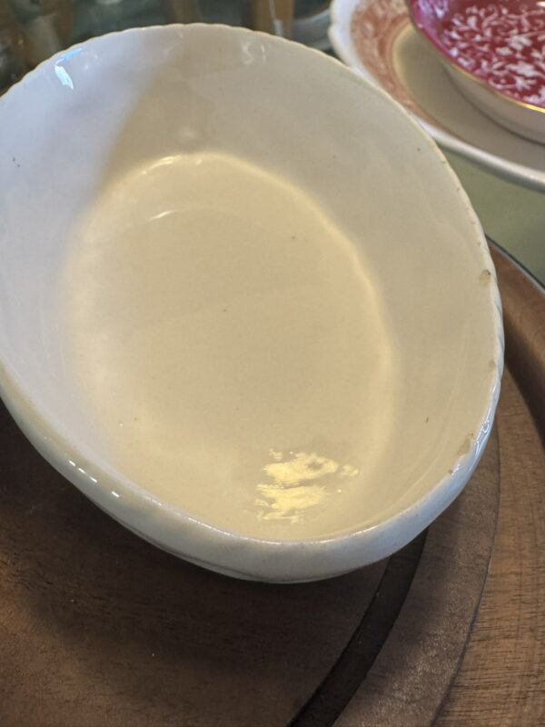 A white, oval Woulind Chile Serving Dish with a slightly uneven rim and minor chips on the right edge sits empty and glossy on a round wooden tray, with part of a red and white patterned plate in the background.