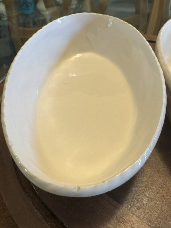 The Woulind Chile Serving Dish is a handmade, oval white ceramic bowl with a slightly uneven rim and glossy finish resting on wood. Subtle edge imperfections give this artisanal serving dish its rustic character.