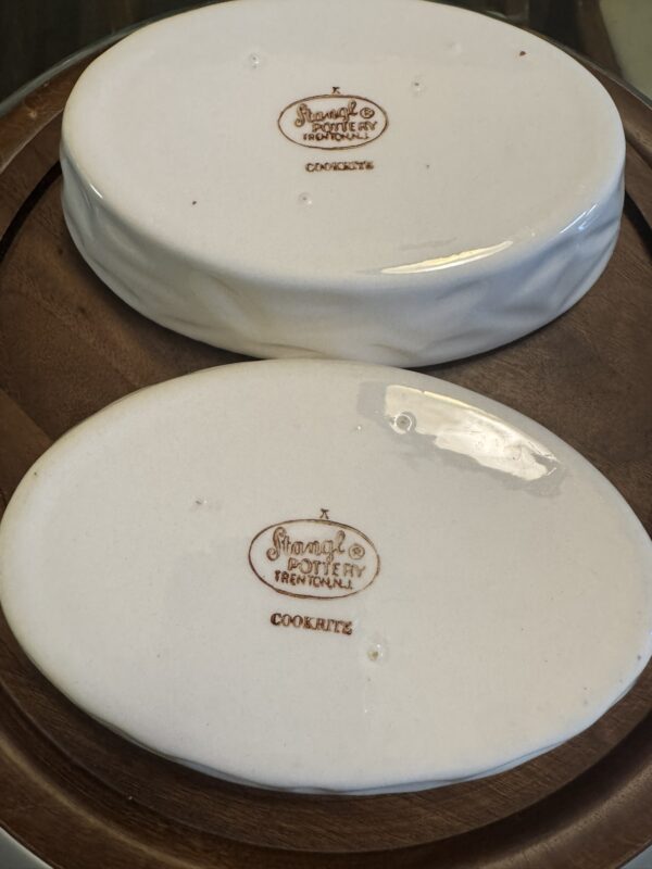 Two oval Woulind Chile Serving Dishes, white ceramic with a glossy finish and small indentations, rest upside down on brown wood. Their stamped marks in brown read