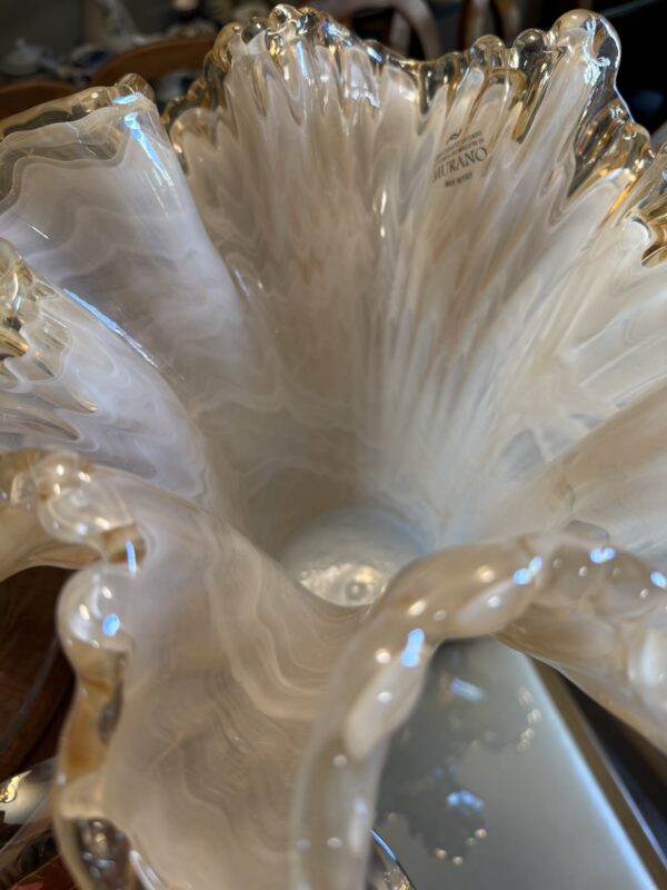 A close-up of the Woulind Chile Serving Dish shows its ornate cream surface with glossy, floral-inspired swirls and ruffled, gold-rimmed edges. Warm light highlights the intricate design and a label on the inner rim.
