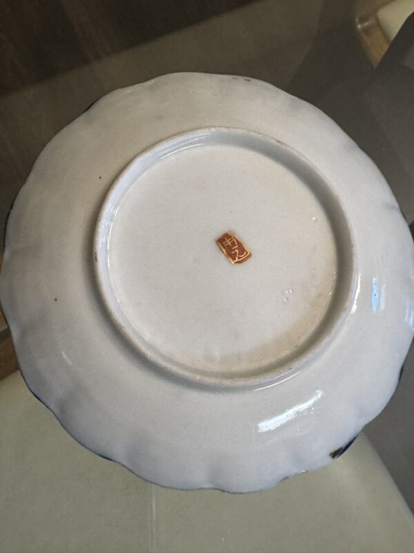 The image shows the underside of the white Woulind Chile Serving Dish with scalloped edges. A small rectangular red label with a gold Asian character is in the center. The dish is placed on a glass surface.