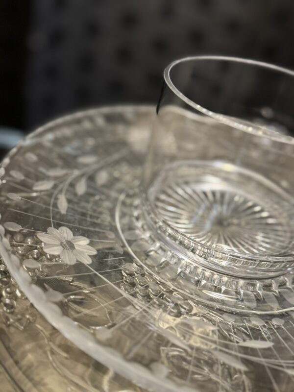 A clear glass cup sits on a Woulind Chile Serving Dish adorned with intricate floral and grape patterns. The softly blurred background highlights the elegant textures of the Woulind dish and glassware.