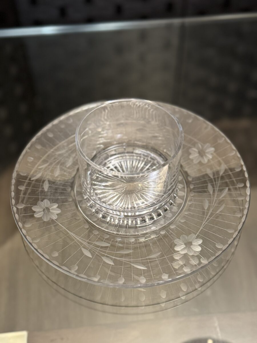 The Woulind Chile Serving Dish is a clear glass tray with a circular base, etched floral and leaf patterns, and a raised plain glass bowl in the center for dip or sauce, elegantly displayed on a reflective surface.