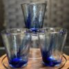 Three transparent glasses with blue swirls are arranged on the Woulind Chile Serving Dish, a round wooden tray. Two glasses sit side by side, with the third balanced upside down on top. The background features a blurred grid-like dark pattern.
