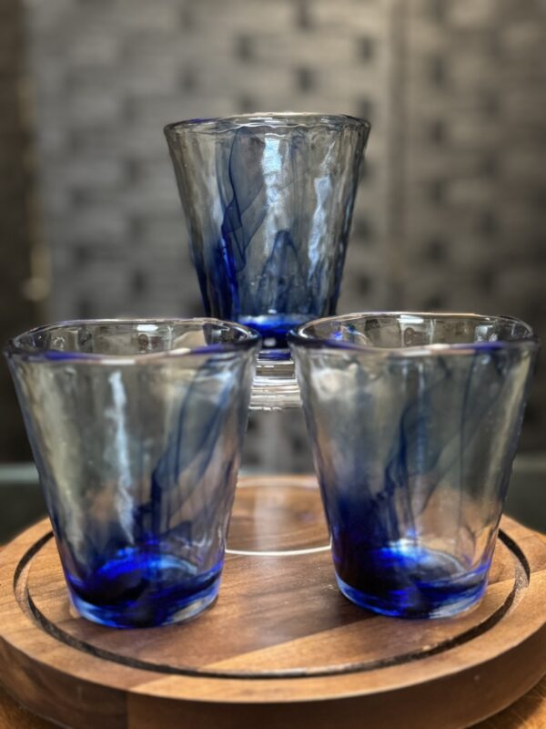 Three transparent glasses with blue swirls are arranged on the Woulind Chile Serving Dish, a round wooden tray. Two glasses sit side by side, with the third balanced upside down on top. The background features a blurred grid-like dark pattern.