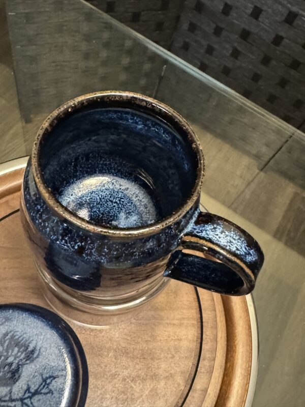 A dark blue ceramic mug with a speckled glaze and matching lid sits on a round wooden tray. The overhead photo reveals a Woulind Chile Serving Dish nearby, set on a glass tabletop with a woven chair in the background.