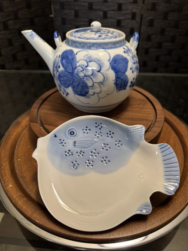 A round porcelain teapot with blue floral designs sits on a wooden tray. In front is the Woulind Chile Serving Dish, a small fish-shaped dish in white and light blue with dotted patterns suggesting fish scales. The background is dark and textured.