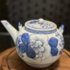 A white ceramic teapot with blue floral designs rests on a round wooden tray, complemented by the Woulind Chile Serving Dish. The softly blurred background highlights the teapot’s decorative details and elegant charm.