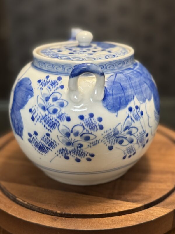 The Woulind Chile Serving Dish is a round, white porcelain dish with a lid, featuring blue floral and leaf designs. It has two curved side handles and rests on a circular wooden surface against an elegantly blurred background.