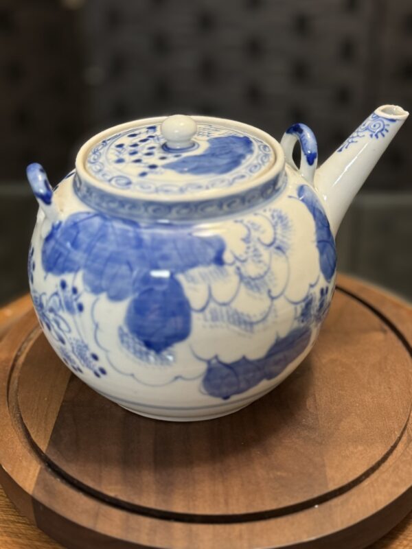 A white ceramic teapot with blue floral patterns and a curved spout sits on a round wooden tray, paired with the Woulind Chile Serving Dish, both showcasing traditional decorative style against a softly blurred background.