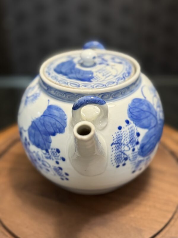 A round white ceramic teapot with blue floral and leaf designs sits beside a Woulind Chile Serving Dish on a wooden surface. The teapot features a central spout, circular handle, lid, and is set against a dark, softly blurred background.