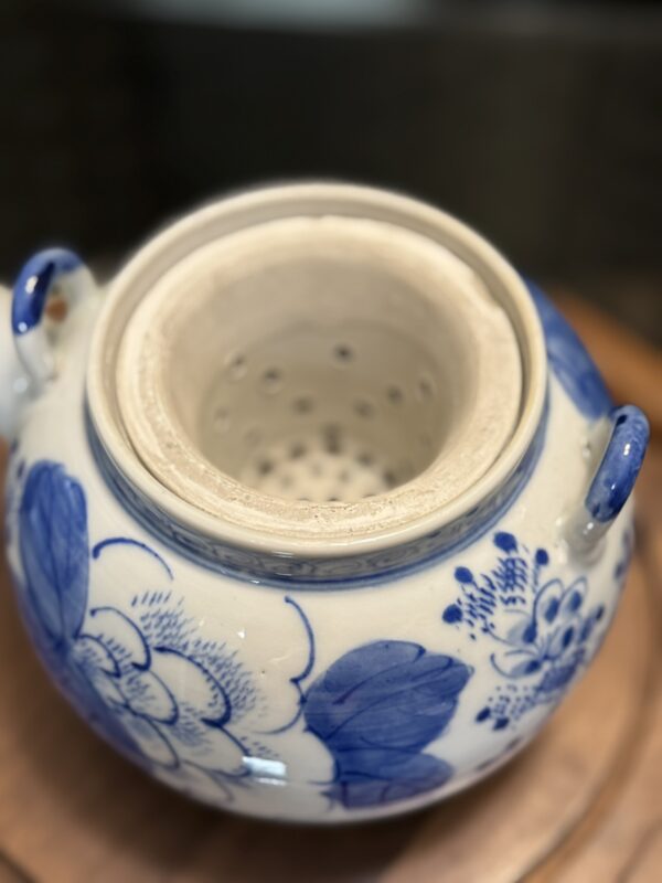 The Woulind Chile Serving Dish features two handles, a wide opening, and an inner chamber with small holes. Its white ceramic body is adorned with large blue flowers and leaves, making it perfect for serving on any wooden surface.