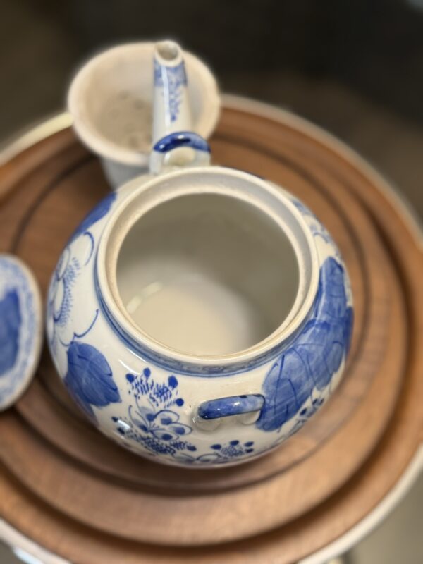 A blue and white ceramic teapot with floral patterns rests on a round wooden tray beside a Woulind Chile Serving Dish. The teapot's lid is off, showing its empty interior, while a matching cup and lid appear in the blurred background.