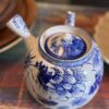 A round ceramic teapot with blue floral patterns sits on the Woulind Chile Serving Dish, a multicolored woven piece. The teapot and handle both feature blue designs, and a white plate is partially visible nearby.