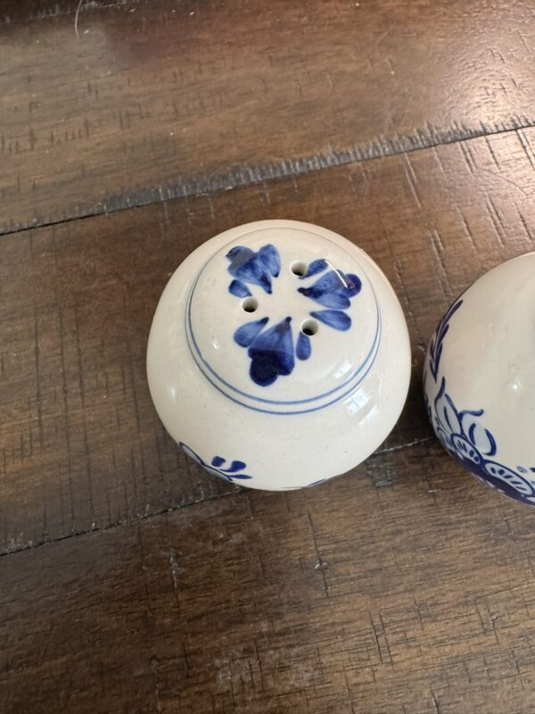 A round, cream ceramic shaker with blue floral patterns and a thin blue rim sits on wood next to the Woulind Chile Serving Dish. The shaker has holes on top, and part of its matching pair appears on the right.