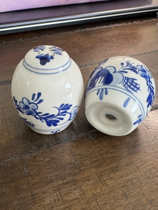 Two Woulind Chile Serving Dishes with blue floral designs are on a wooden surface. One stands upright, lid displayed; the other lies on its side, showing a hole in the base. A pink and metallic edge is visible in the blurred background.