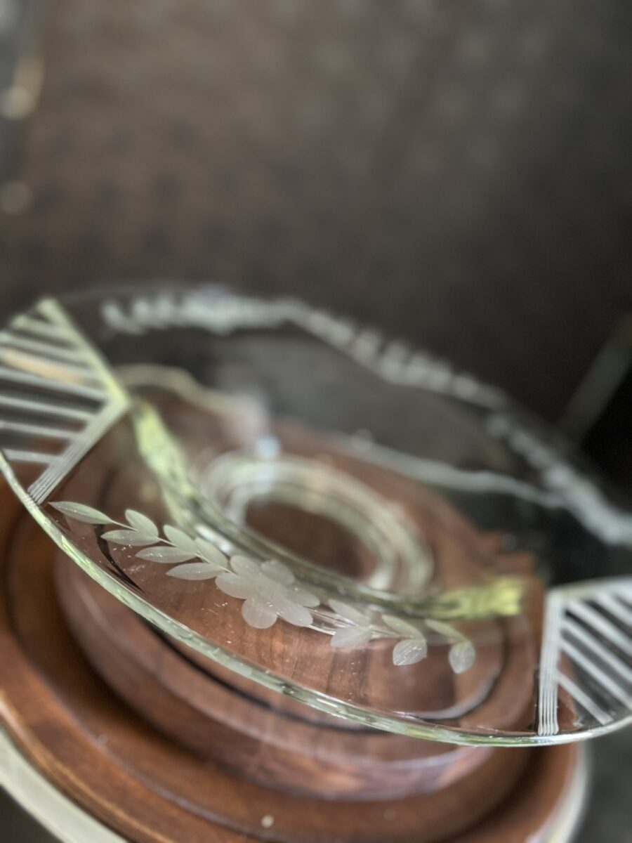 A close-up of the Woulind Chile Serving Dish shows its clear, round design with etched leaf and line patterns around the rim, a scalloped edge, and a wooden stand, photographed at an angle against a dark, blurred background.