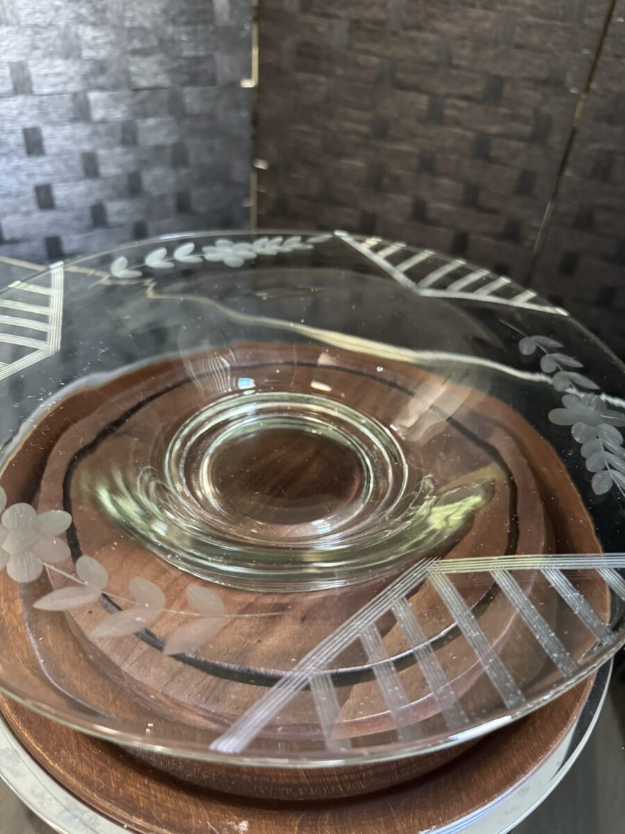 The Woulind Chile Serving Dish features clear glass etched with leaves and diagonal lines, set on a round wooden base. Against a dark woven-texture background, reflected light emphasizes the dish’s transparency.