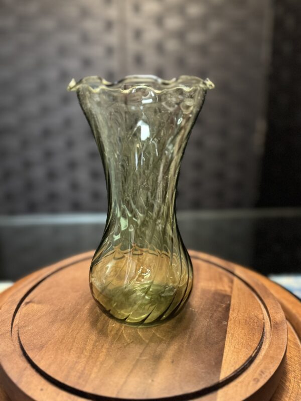A tall, green glass vase with a fluted rim and swirling pattern stands on a wooden tray beside the elegant Woulind Chile Serving Dish, their delicate textures highlighted against a dark background.