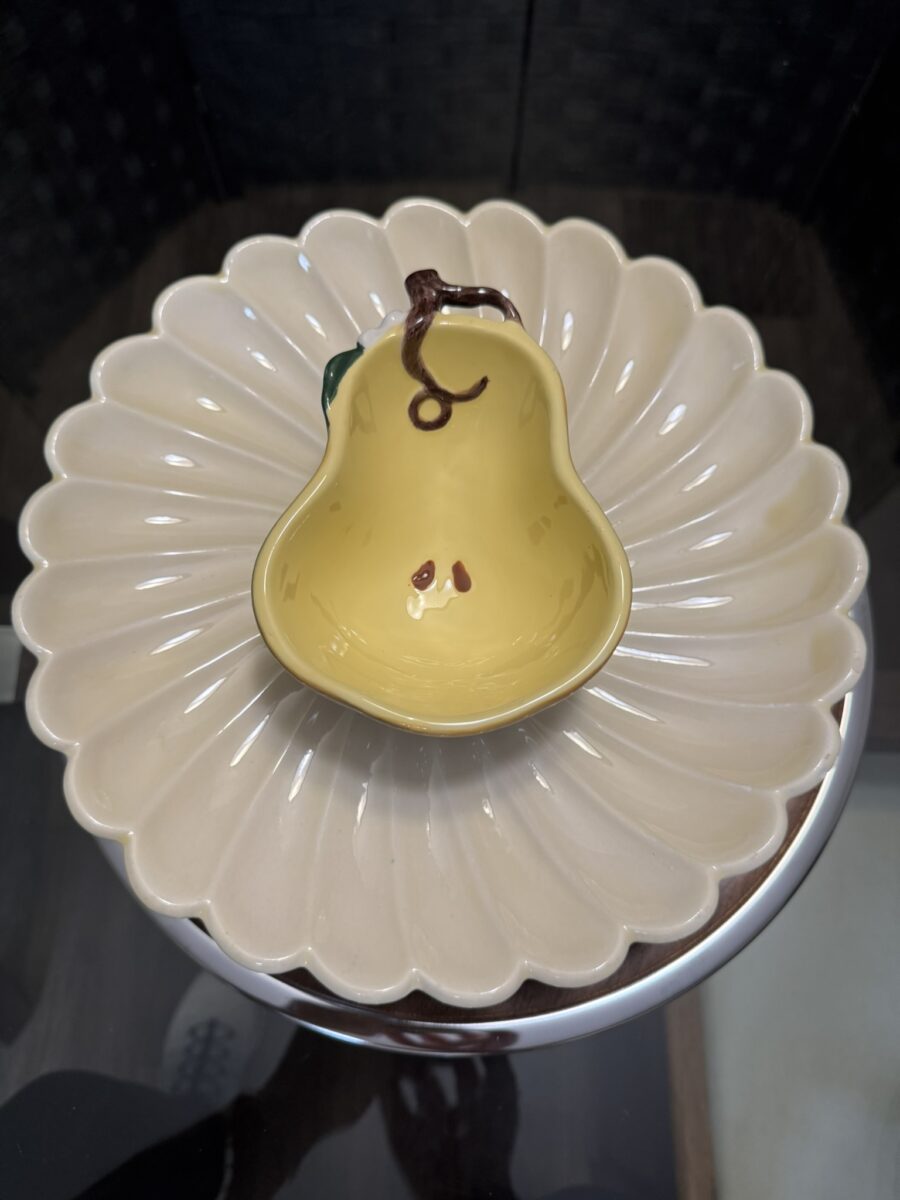 The Woulind Chile Serving Dish, a small yellow ceramic piece with a brown stem and green leaf, sits centered on a round cream plate with scalloped edges, creating a floral display on a reflective glass surface.