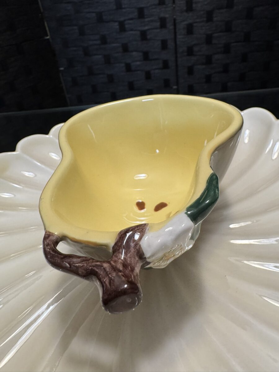 The Woulind Chile Serving Dish, a yellow ceramic corn cob with a brown handle and green leaf detail, sits on a cream petal-shaped plate against a dark textured background.