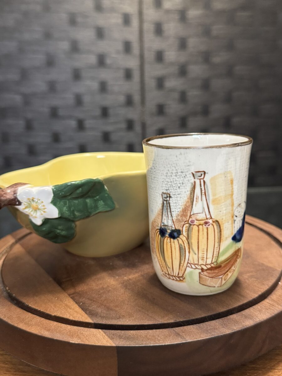 A small ceramic cup with bottle, grape, and fruit illustrations sits on a round wooden surface beside the Woulind Chile Serving Dish, which features a yellow hue, flower-shaped handle, and green leaves against a dark textured grid background.