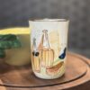 A ceramic cup with hand-painted designs of bottles, cheese, and olives rests on a round wooden board next to the Woulind Chile Serving Dish—a yellow bowl featuring a sculpted green leaf—set against a softly blurred, cozy background.