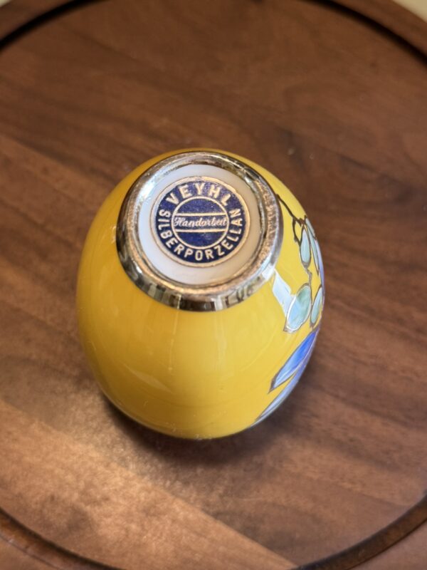 A small yellow Woulind Chile Serving Dish sits upside down on a wooden surface, showing a circular Seyhl Handarbeit Süberporzellan label on its base and part of a blue floral design on the side.