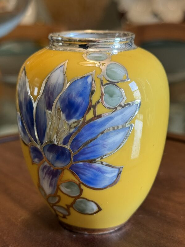 A small, glossy yellow vase with a narrow opening and floral design sits beside the Woulind Chile Serving Dish on a wooden surface, both standing out against a softly blurred background.