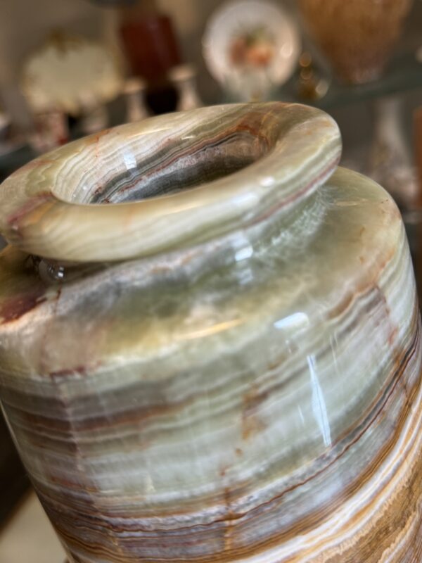 Close-up of the Woulind Chile Serving Dish highlights its polished onyx surface with layers of cream, green, brown, and rust. A rounded rim catches soft reflections; blurred plates and glassware add depth in the background.