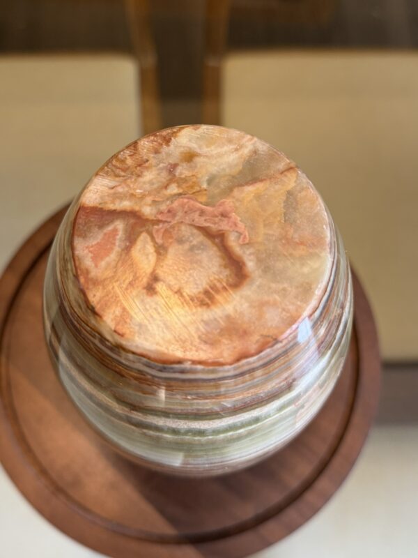 A top-down view of the Woulind Chile Serving Dish, a round polished stone with earthy pink, beige, and brown swirls. It sits on a circular wooden base, showcasing its textured surface and natural shine.