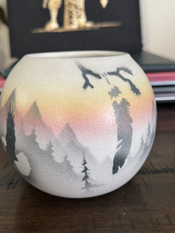 The Woulind Chile Serving Dish is a round ceramic piece with a matte finish, featuring a hand-painted landscape of pine trees and distant mountains. The sky fades from yellow to orange and pink, evoking the colors of sunrise or sunset.